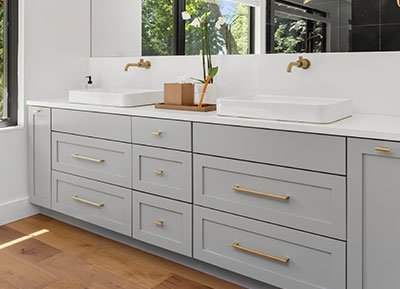 A modern bathroom with light gray double vanity featuring gold hardware, two white rectangular sinks, and gold wall-mounted faucets. A white orchid in a wooden pot sits between the sinks. The countertop is white, and there is a window view of greenery in the background—perfectly curated by top home remodeling contractors.
