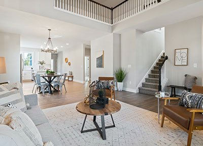 A modern, open-concept living room with neutral tones, crafted by top-tier home company professionals. The room features a cozy sofa, wooden coffee table, and chairs. A staircase with a railing leads upstairs. The dining area in the background has a round table with chairs and a chandelier. Decor includes plants and wall art.