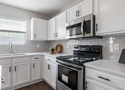 Modern kitchen with white cabinets, stainless steel appliances including a microwave and oven, light gray tile backsplash, and dark wood flooring. A window above the sink allows natural light to brighten the space. Decor includes a wicker basket and a small plant—courtesy of top home remodeling contractors.