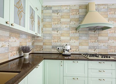 A modern corner kitchen with light green cabinets and dark countertops. The backsplash features tiles in various pastel colors and patterns. Recently renovated by top kitchen remodel contractors, it includes a stove with a range hood and a small appliance on the counter next to dried lavender.