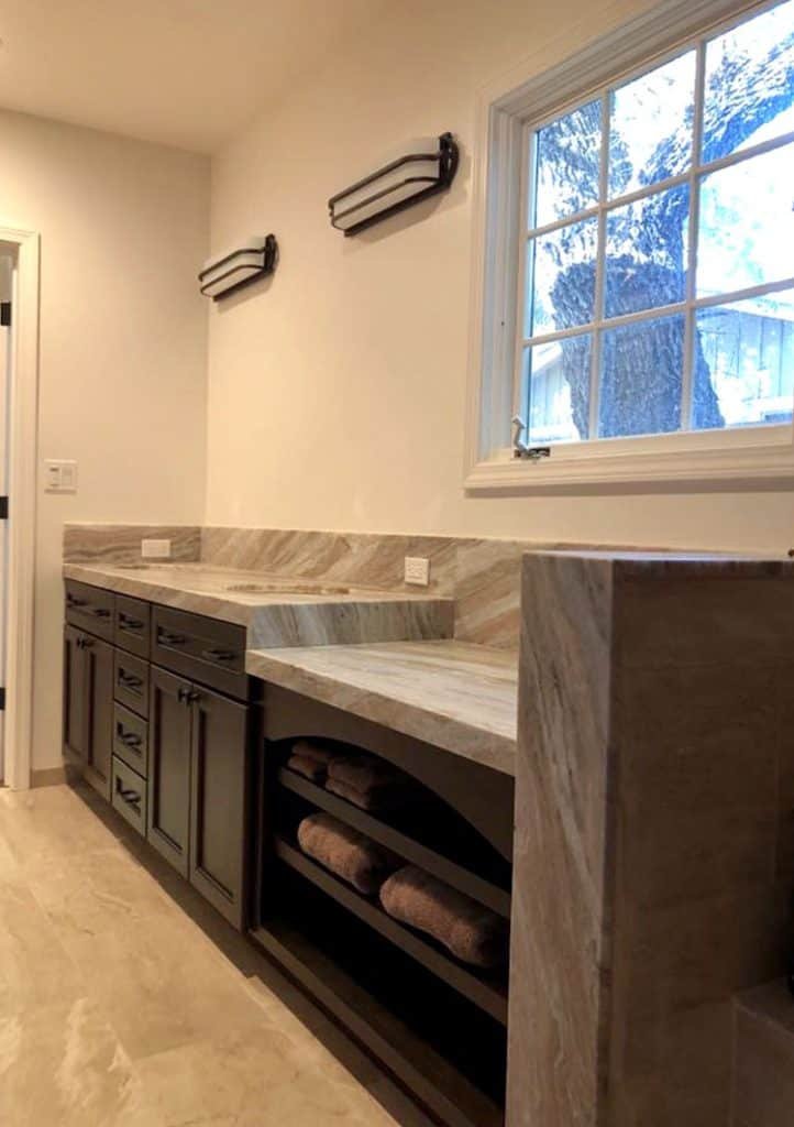 Modern bathroom with light wooden cabinets and marble countertops. Open shelving holds neatly folded towels. Two wall-mounted lights are above the vanity, and a large window allows natural light to enter. The minimalist design emphasizes functionality and style, perfect for those planning a renovation.