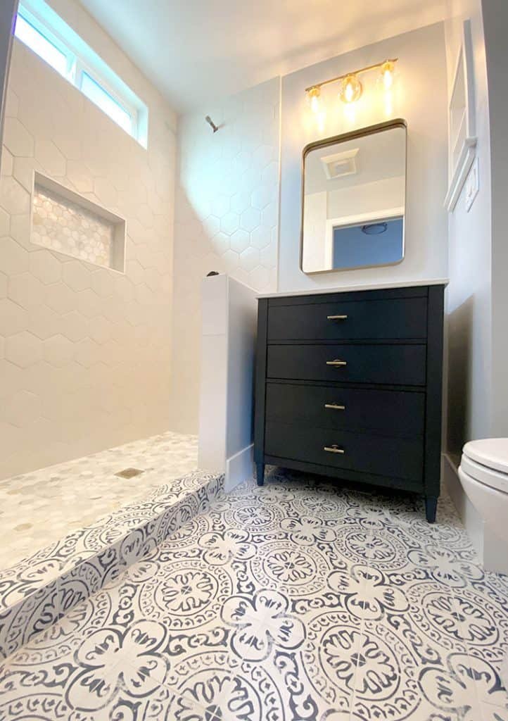 A modern bathroom designed by top home remodeling contractors features patterned tile flooring, a black vanity with three drawers and a rectangular mirror above it, and a walk-in shower with white hexagonal tiles and a small recessed shelf. The shower has a half-wall separating it from the vanity area.