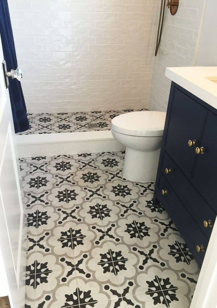 A small bathroom with white subway tile walls and patterned black and white floor tiles, newly finished by a home company. It features a white toilet, dark blue vanity with gold knobs, and a shower curtain likely hiding the shower area in the background.