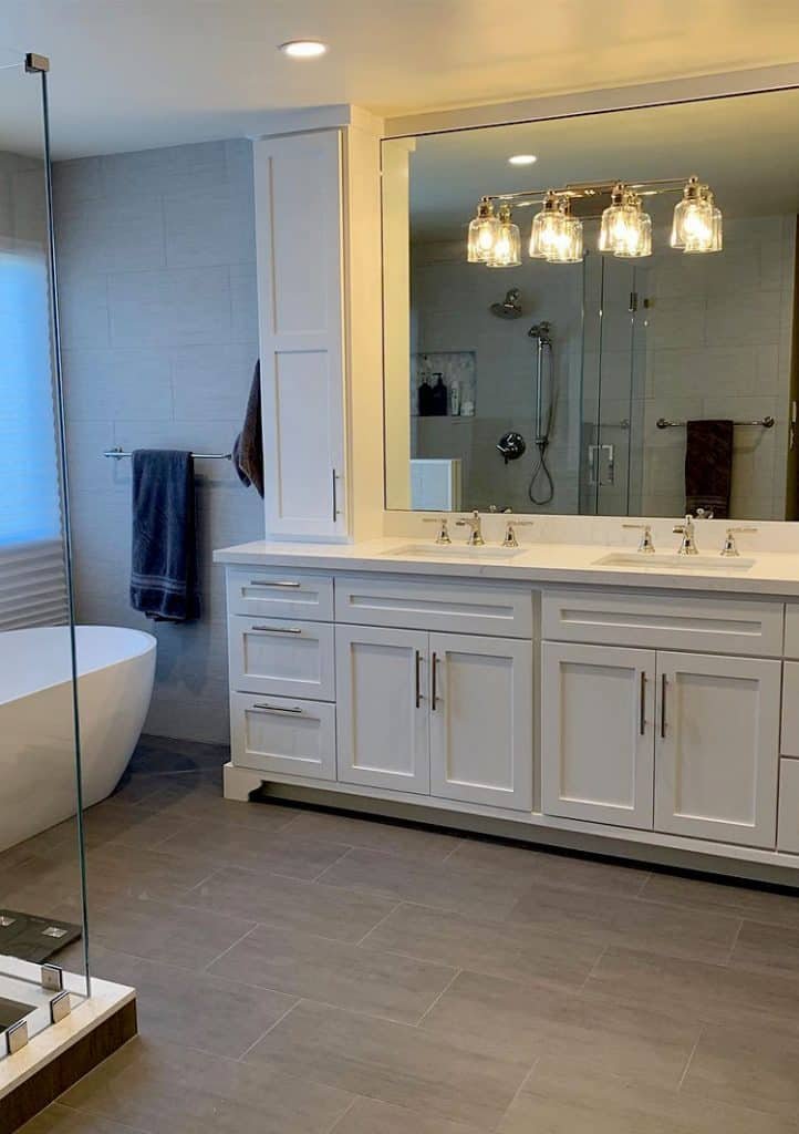 A modern bathroom with a large mirror above a double sink vanity featuring white cabinets and silver handles. There's a freestanding bathtub near the window, a towel hanging on a rod, and a glass-enclosed shower with a rainfall showerhead in the background, all reflecting top-tier renovation standards by our home company.