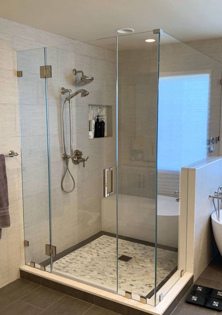 A modern bathroom by home remodeling contractors features a glass-enclosed shower with a rainfall showerhead and built-in wall niche for toiletries. Adjacent to the shower is a white bathtub with a chrome faucet. The bathroom boasts beige tiles and a towel hanging on the wall, showcasing expert craftsmanship.