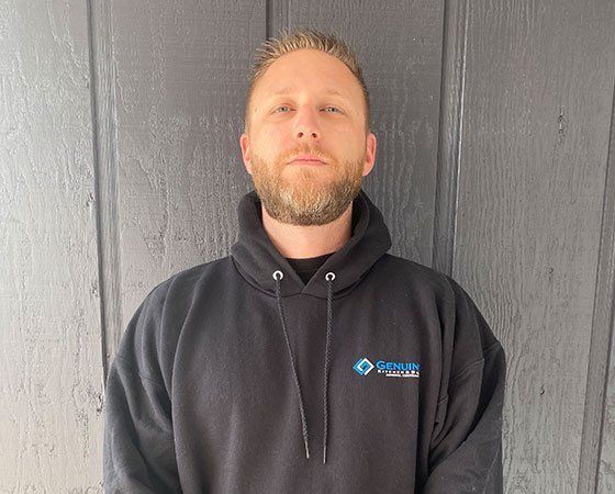 A person with short hair and a beard stands in front of a wooden wall, likely the work of skilled home remodeling contractors. They are wearing a black hoodie with the logo and text "GENUIN" above smaller, unreadable text on the upper left side. The person has a neutral expression and is looking straight at the camera.