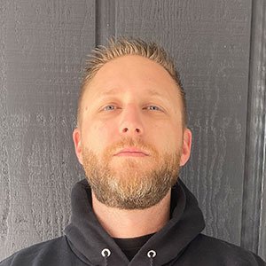 A man with short, light brown hair and a trimmed beard is standing against a dark-colored wooden wall, wearing a black hoodie. He has a neutral expression and is looking directly at the camera, resembling an experienced home remodeling contractor ready for his next project.