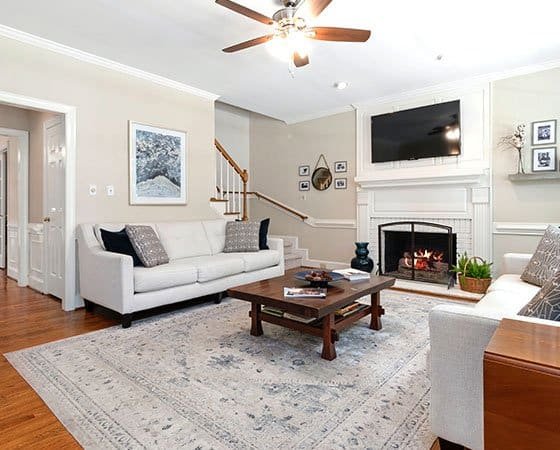A cozy living room features a white sofa with patterned cushions, a wooden coffee table on a light-colored rug, and a fireplace with a TV mounted above it. A second sofa complements the space, while the walls are adorned with framed art. Expertly finished by our home company, the ceiling fan completes the look.