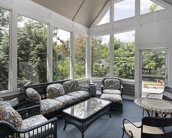 A sunroom with large windows and a glass door provides a view of lush greenery outside. The room, furnished with a wicker sofa set featuring floral cushions, a glass-top coffee table, and a small side table, showcases the impeccable work of home remodeling contractors. The high vaulted ceiling creates an airy feel.