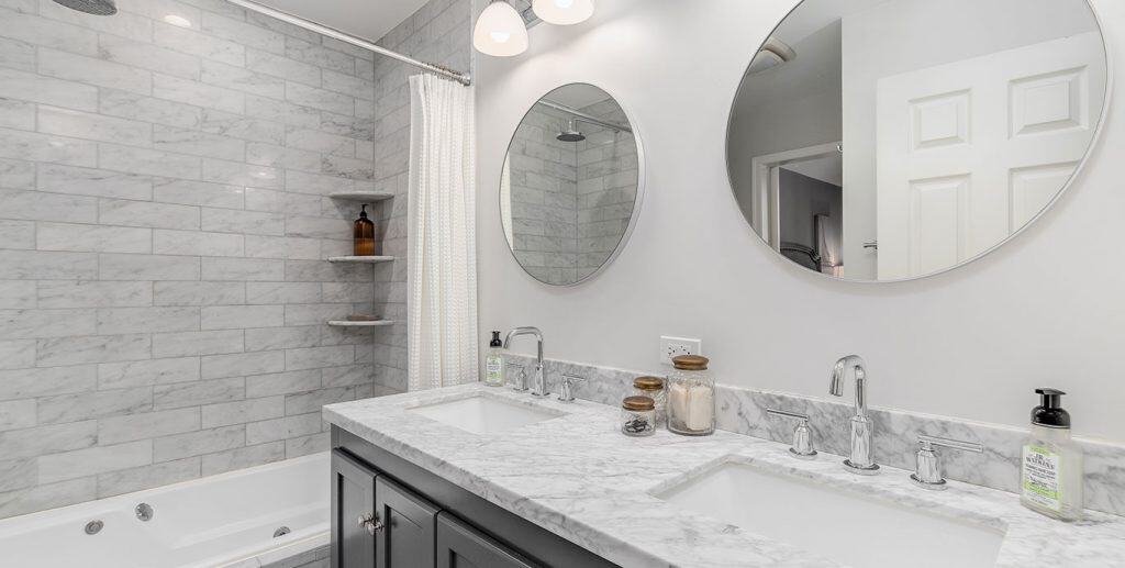 A modern bathroom, perfect for any renovation project, features a marble countertop with dual sinks. Above the sinks are two circular mirrors and light fixtures. The background includes a tiled shower area with a white curtain, built-in shelves, and a closed door on the right side.