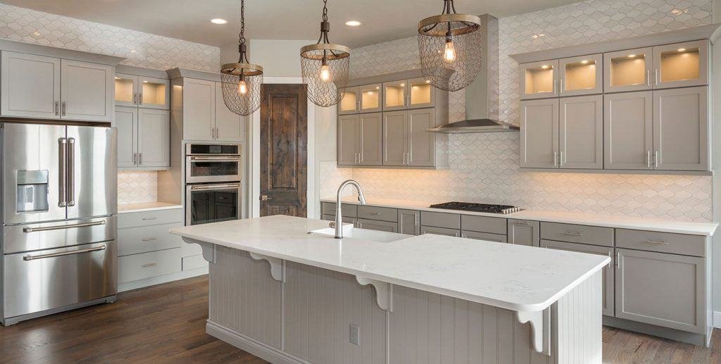 Modern kitchen featuring a large island with a white countertop and built-in sink. Grey cabinets and stainless steel appliances, including a double oven and refrigerator, are complemented by three pendant lights and hexagonal tile backsplash. Wood floors complete the space, courtesy of top-notch kitchen remodel contractors.
