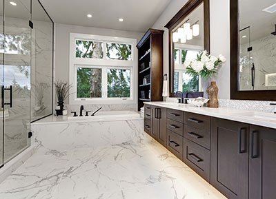 Luxurious bathroom with white marble flooring and countertops, perfected by our home company's expert renovation team. Features a large soaking tub by the window, a glass-enclosed shower, dark wood cabinetry, twin sinks with black fixtures, and a large mirror. A vase with white flowers decorates the countertop.