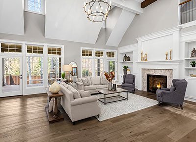 A spacious living room with a high, vaulted ceiling and large windows letting in natural light. It features a white sectional sofa, two gray armchairs, a coffee table, and a lit fireplace. Renovation by expert home remodeling contractors ensured the decor includes various ornaments and a chandelier hanging from the ceiling.
