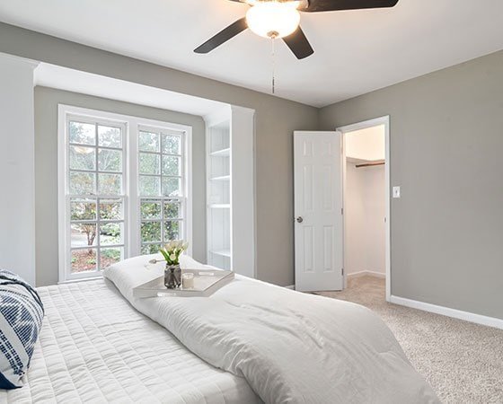 A minimalist bedroom with light gray walls, a large window framed by white built-in shelves, a bed with white bedding, decorative pillows, and a tray of flowers on the bed. A ceiling fan with a light hangs above, courtesy of top home remodeling contractors. An open door leads to the closet.