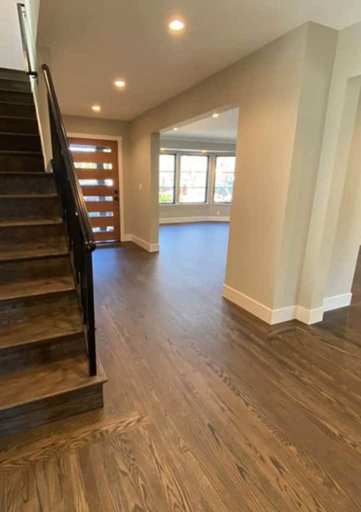 A modern home interior by top home remodeling contractors features a wooden staircase with a metal railing to the left. The entryway is well-lit with recessed lighting, and the floor is dark hardwood. The integrated living spaces have large windows that invite natural light.