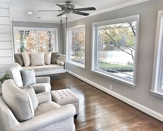 A sunlit room with large windows overlooks a serene river and trees. It features light grey walls, a white ceiling fan, and wooden flooring. Thanks to skilled home remodeling contractors, the space boasts comfortable beige seating, including a cushioned armchair with a footrest and a built-in window seat with pillows.