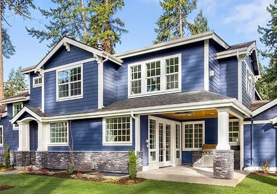 A two-story house with blue siding and white trim stands surrounded by tall trees. Thanks to skilled home remodeling contractors, the house features multiple large windows, a covered front porch with white columns, and a stone accent on the lower half of the exterior. The yard has neatly manicured grass and shrubs.