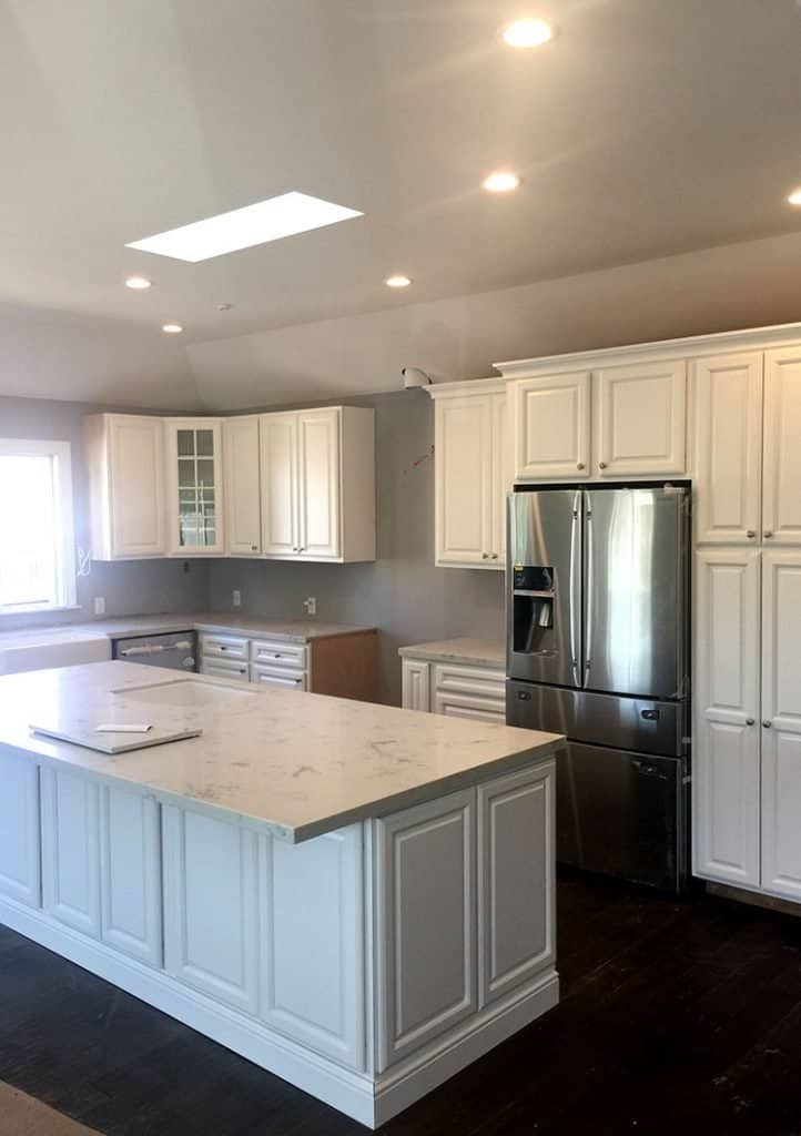 A spacious, modern kitchen with white cabinets, a large marble-topped island, and stainless steel appliances, including a double-door refrigerator. The room has recessed lighting and a skylight, providing ample natural light—a perfect showcase of home remodeling contractors' expertise.