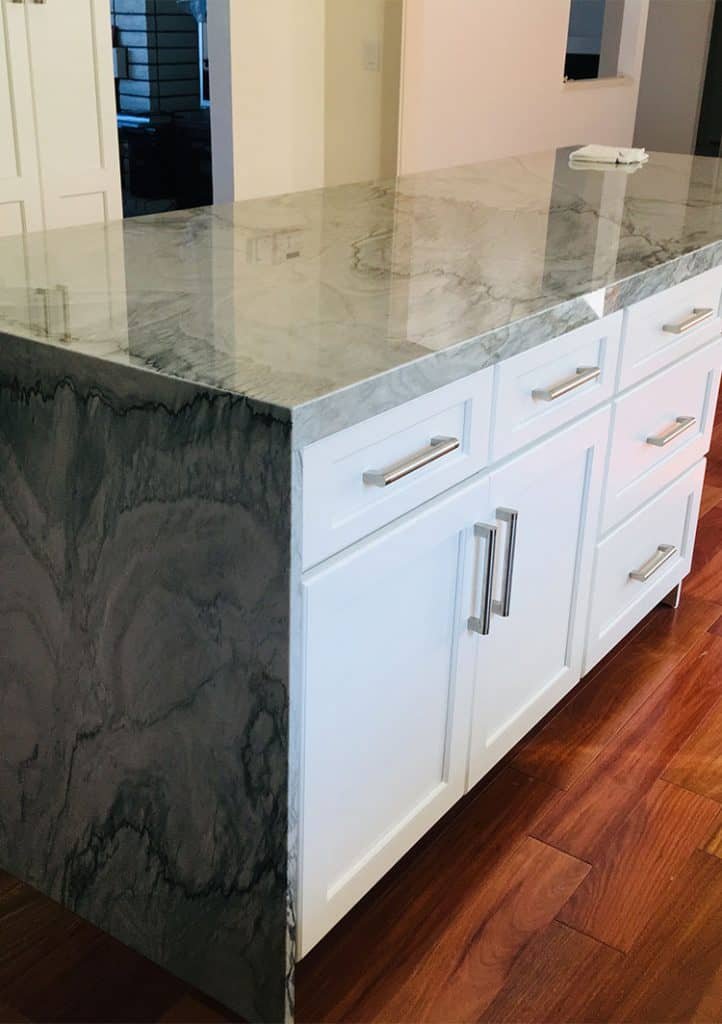 A kitchen island with a polished gray marble countertop and white cabinetry with silver handles. The side of the island features a matching gray marble surface. The wooden floor adds warmth, and a small part of the surrounding kitchen is visible—a testament to quality work by top-notch kitchen remodel contractors.
