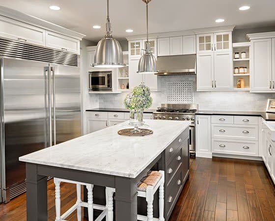 Modern kitchen with stainless steel appliances, including a large refrigerator and oven. White cabinets with glass-paneled upper sections line the walls. A gray island with a white marble countertop stands centrally, under two pendant lights, with flowers in a vase—courtesy of expert home remodeling contractors.