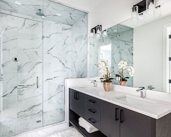 A modern bathroom featuring a spacious glass shower with marbled tile walls and a large vanity with a white countertop and dual sinks. The vanity, crafted by expert home remodeling contractors, has dark cabinetry adorned with potted white orchids and a large mirror above.