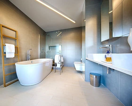 A modern, spacious bathroom with a freestanding bathtub, a wall-mounted toilet, a double sink vanity with a large mirror, and a glass-enclosed shower. The room features light-colored tile flooring, gray walls, a wooden towel ladder, and sleek contemporary fixtures—perfect inspiration for your next home remodeling project.