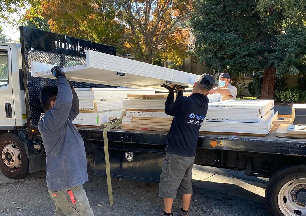 Three people from a home company are unloading white door frames from a flatbed truck on a sunny day. Two men in the foreground lift a large frame, while a third person in the background manages the remaining frames on the truck. They are near a tree with autumn-colored leaves, likely for a renovation project.