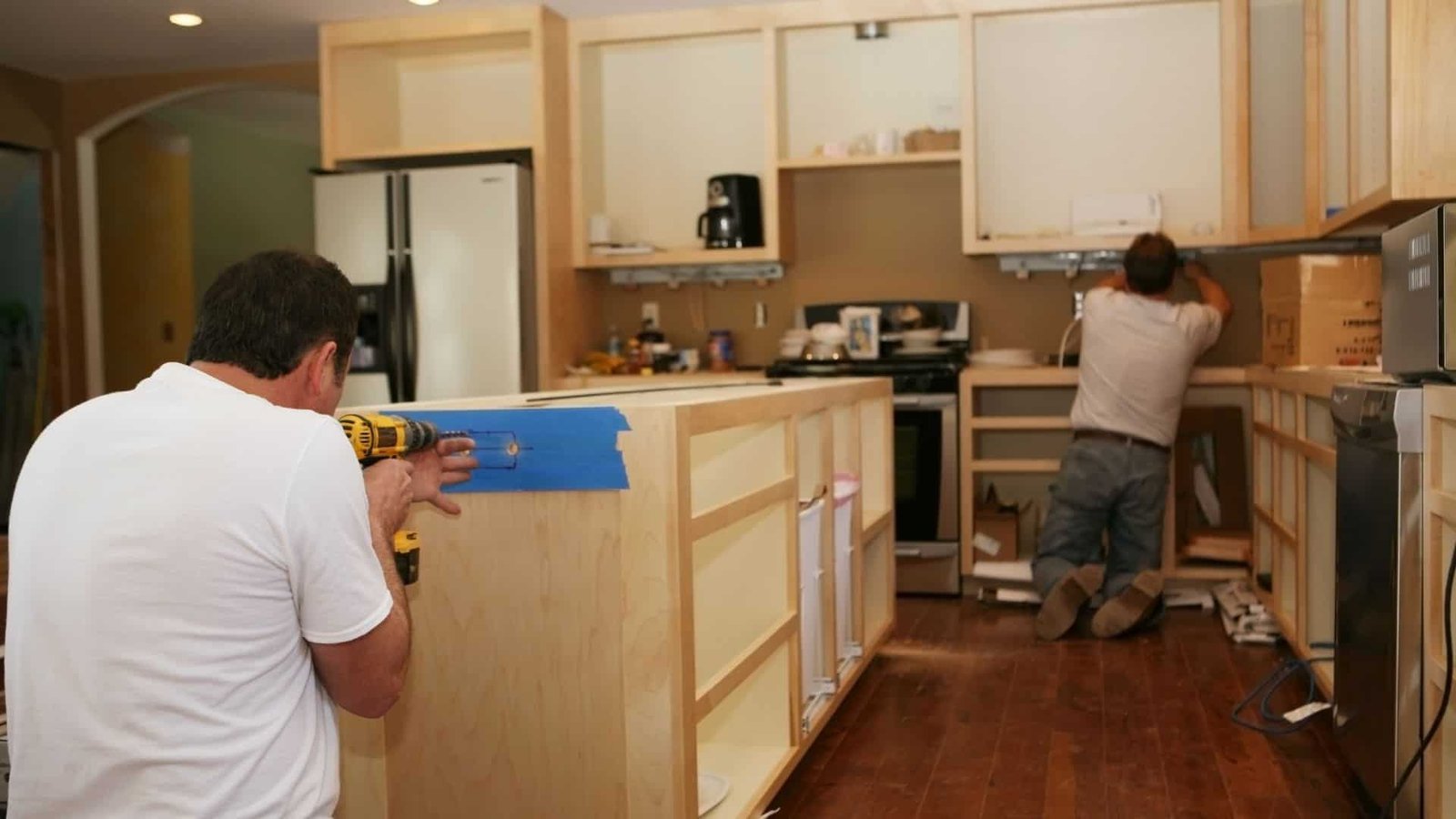 Two men, experienced kitchen remodel contractors, are working on a kitchen renovation. One man is drilling into a wooden cabinet with a blue tape guide, while the other is adjusting something on the kitchen wall near the stove. The kitchen is partially completed with unfinished cabinetry.
