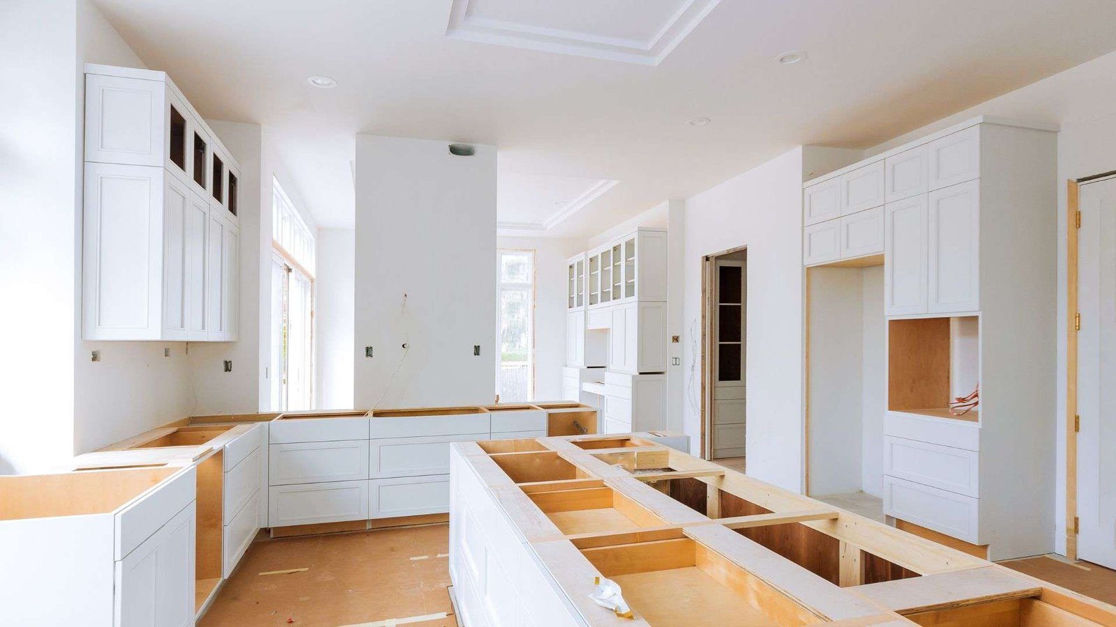 A bright, partially constructed kitchen with white cabinets and countertops not yet installed. The room has ample natural light coming through large windows. Cabinet frames and some drawers are visible, with tools and construction materials placed around by the kitchen remodel contractors.