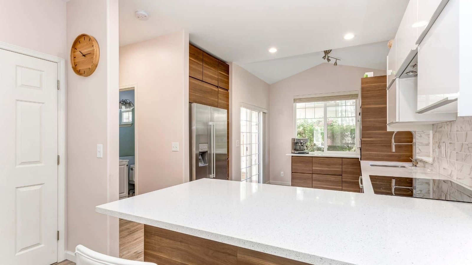 A modern kitchen with white countertops and wooden cabinets. It features a fridge, an oven, and a stove. A clock hangs on the wall near a doorway, and large windows allow natural light to flood the space. Recently renovated by home remodeling contractors, it includes a white door to the left and barstool seating.