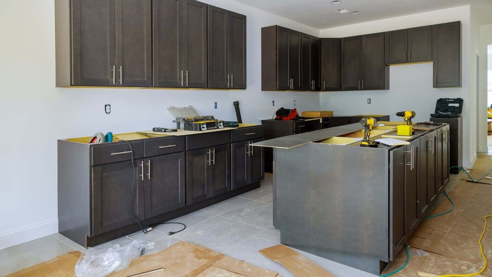 A modern kitchen under renovation with dark gray cabinets and drawers installed. The countertop surfaces are not yet placed. Tools, materials, and boxes are scattered across the unfinished floor while wiring and outlets are visible on the walls.