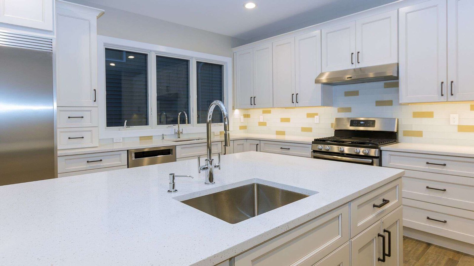 Modern kitchen with white cabinets and a large island featuring a built-in sink and chrome faucets. The room includes stainless steel appliances, including a refrigerator and stove, and a window over a secondary sink. The backsplash is a mix of white and yellow tiles, all part of our home company's expert renovation services.