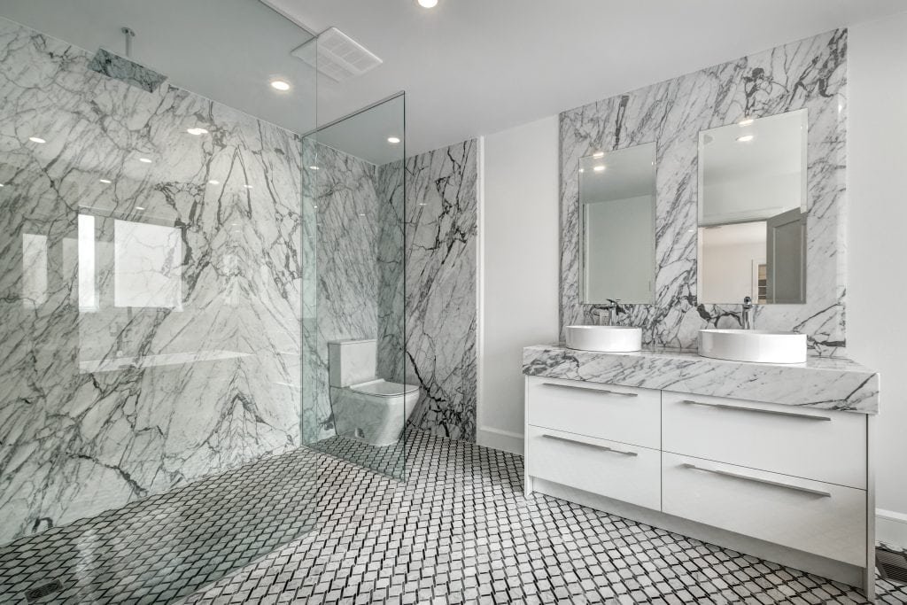 A recent renovation of this modern bathroom showcases a large walk-in shower with glass doors and marble walls. Adjacent is a double sink vanity with marble countertops and two mirrors. The floor is adorned with intricate, hexagonal black and white tiles, enhancing the luxurious decor.