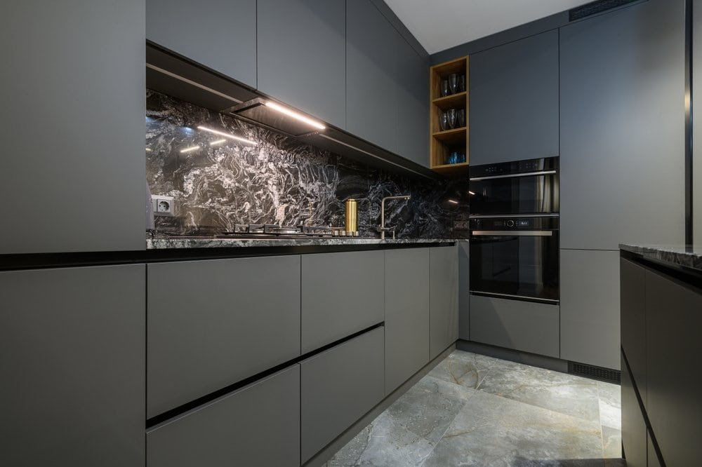 A modern kitchen with dark gray cabinets and a black marble backsplash. The countertops are also black marble. Built-in appliances, including an oven and microwave, are integrated into the cabinetry. The floor features large stone tiles, creating a stunning space any renovation by expert kitchen remodel contractors would achieve.