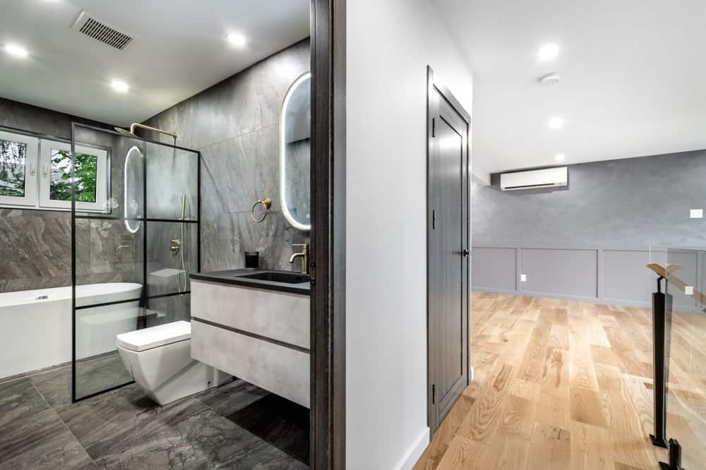 A modern bathroom with grey tiles features a glass-enclosed shower, a wall-mounted toilet, a vanity with a black sink, and an oval mirror. Adjacent to the bathroom is a spacious room with wooden flooring and a wall-mounted air conditioner, masterfully crafted by top home remodeling contractors.