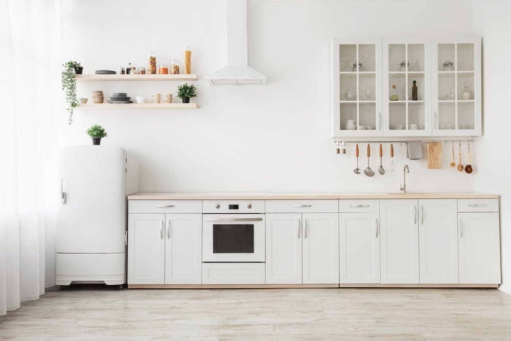 A minimalist kitchen with white cabinets, a stove, and a sink. Shelving holds dishes, jars, and plants. A white fridge is on the left. Natural light flows in from a large window, highlighting the clean, modern design.