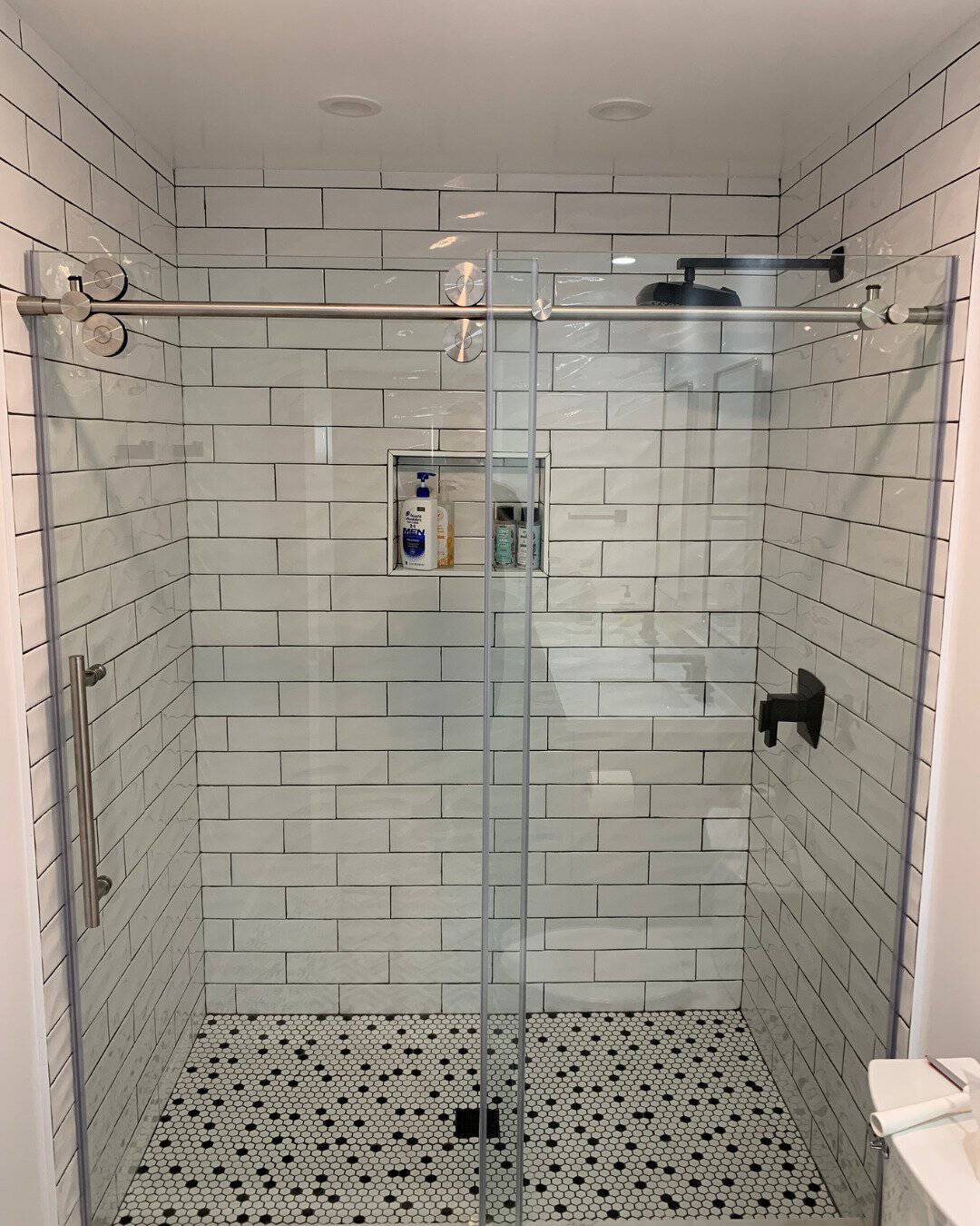 Modern walk-in shower remodel in San Carlos, CA featuring white subway tile walls, black and white penny tile flooring, and a frameless sliding glass door with industrial rollers