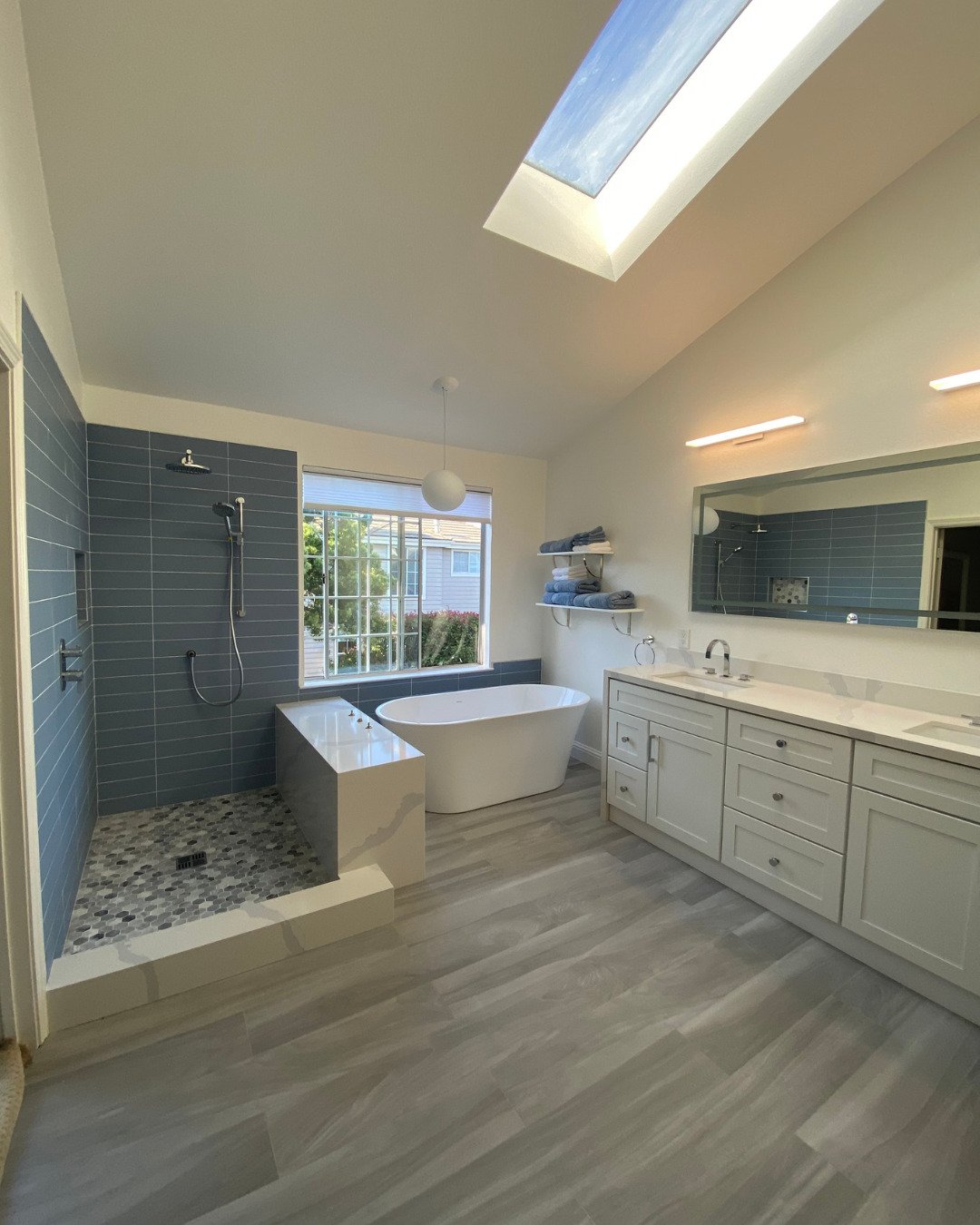 Modern bathroom renovation in San Carlos, CA featuring a vaulted ceiling with a skylight, blue tiled walk-in shower, freestanding soaking tub, and a large white double vanity